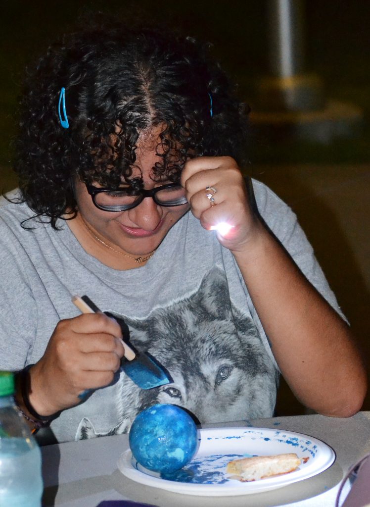 Student paints a small styrofoam ball like a planet during an evening under the stars event.